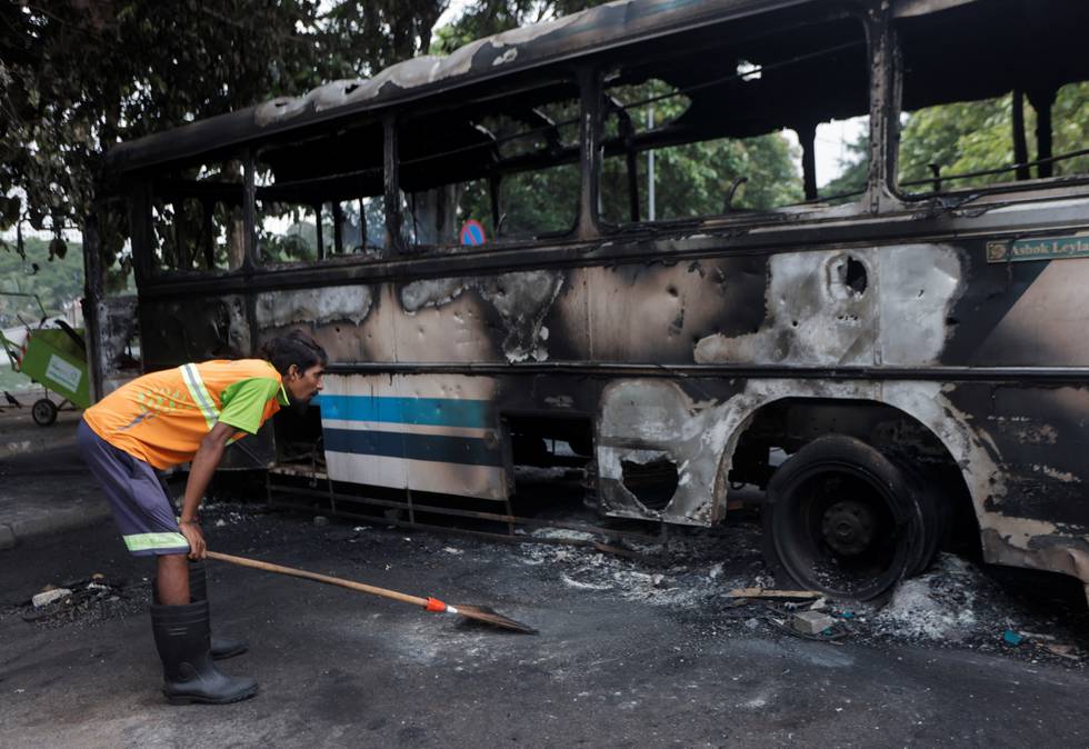 Bildet er av en mann som lener seg framover og tar seg til knærne. Han ser på restene av en utbrent buss. Foto: Dinuka Liyanawatte / Reuters / NTB