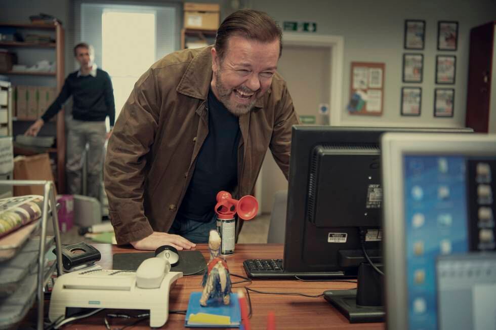 Bildet er av Ricky Gervais som lener seg over en pult og ler. Han ser på en dataskjerm på et kontor. I bakgrunnen ser en mann på ham.