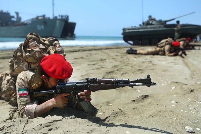 Bildet viser iranske soldater som ligger i sanden. De har kamuflasje for ørkenbruk, og røde luer. De sikter på et mål utenfor bildet. I bakgrunnen er et skip og en stridsvogn.