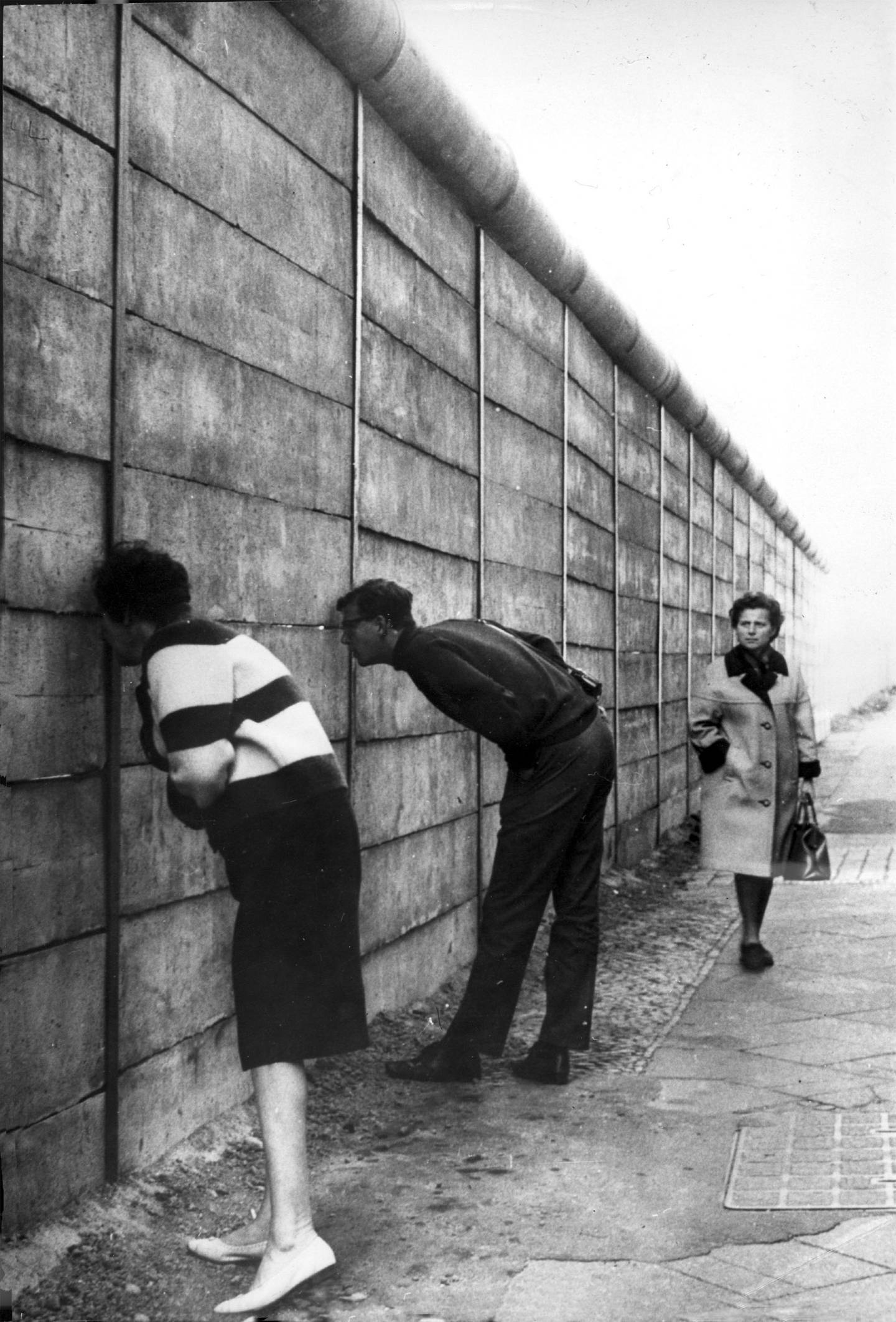 Bildet er av folk i Vest-Berlin som ser inn til Øst-Berlin gjennom sprekker i muren. Den er høy, og har en sylinder på toppen. Det gjør at muren buer ut på toppen, og blir vanskeligere å klatre over. En dame kommer gående i bakgrunnen. Foto: Keystone / Billedsentralen / NTB