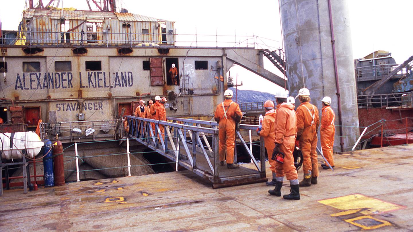 Bildet er av folk i oransje kjeledresser. De står ved en gangbro som spenner fra en brygge og ombord Alexander Kielland.