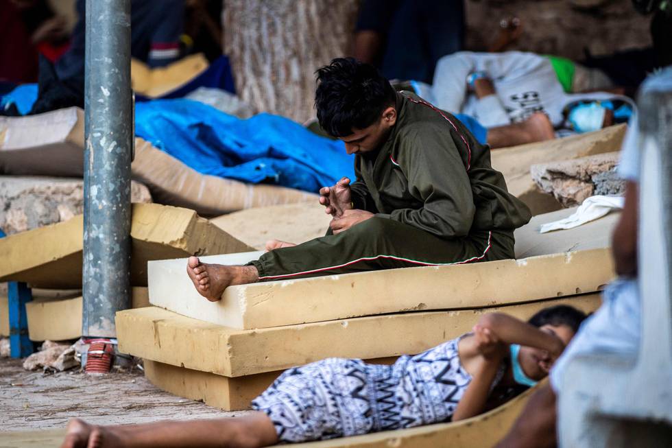 Bildet er av en mann som sitter på en stabel av tre skumgummimadrasser. Det ligger en person på en madrass på siden av ham. Bildet er fra leiren tidligere i juli. Foto: Alessandro Serrano / AFP / NTB