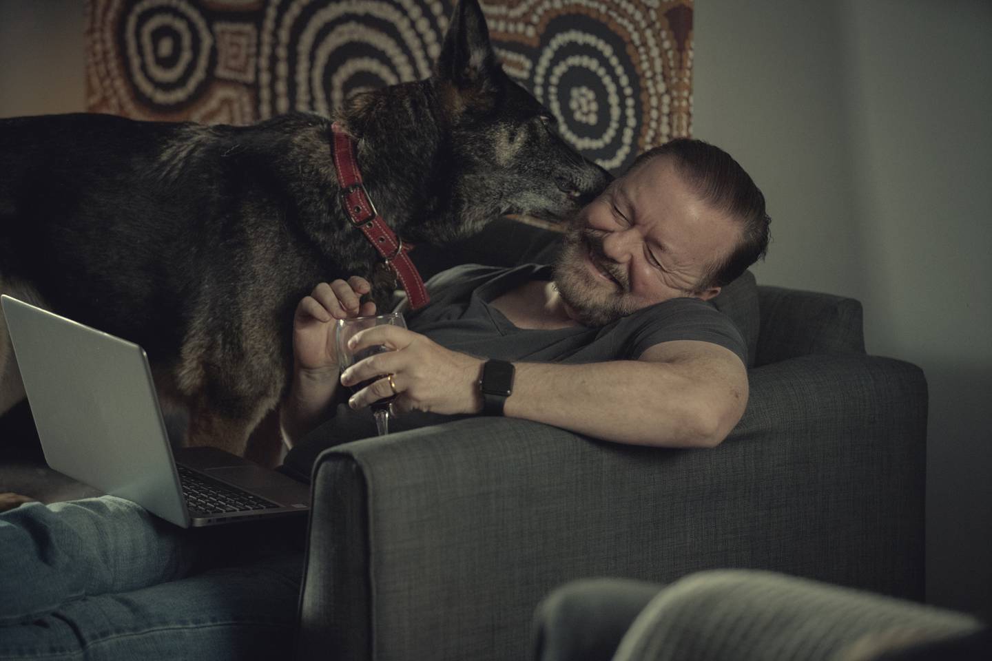 Bildet er av Ricky Gervais som sitter på en sofa med en laptop i fanget. Han smiler med lukkede øyne mens han blir slikket i øret av en stor hund. Det er en schäfer.