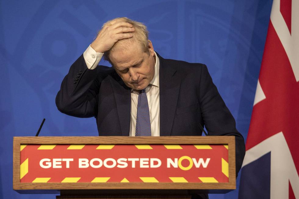 Bildet er av Boris Johnson som står foran en blå vegg og et britisk flagg. På talerstolen foran ham er det en oppfordring til å ta en tredje dose koronavaksine. Johnson har på dress og tar seg til hodet, mens han ser ned.