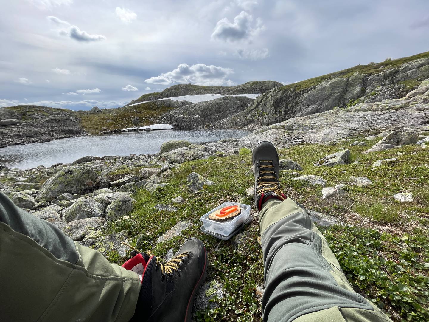 Bildet er av beina til en person som sitter på bakken og strekker dem ut. Personen er på tur på Hardangervidda, og bakken er dekket av lyng og små steiner. Det er et vann lenger framme. Foto: Solveig Vikene / NTB