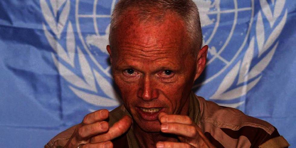 Bildet viser Mood som snakker i en mikrofon foran et FN-flagg. Norske Robert Mood leder FN-observatørene i Syria. De blir i landet selv om de staig blir angrepet.