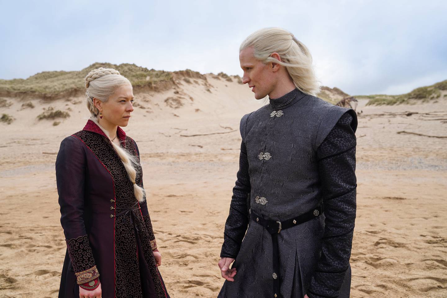 Bildet er av Emma D’Arcy som prinsesse Rhaenyra Targaryen og Matt Smith som prins Daemon Targaryen. De står omgitt av sand. I bakgrunnen er det noen topper med gress. De har på seg mørke antrekk.