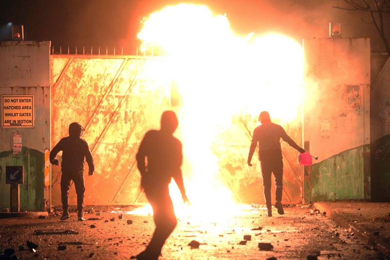 Bildet er av en brann langs en flere meter høy vegg. Tre personer i mørke klær har ryggen mot kamera. En av de holder en beholder. Det ble kastet flere brannbomber over veggen.