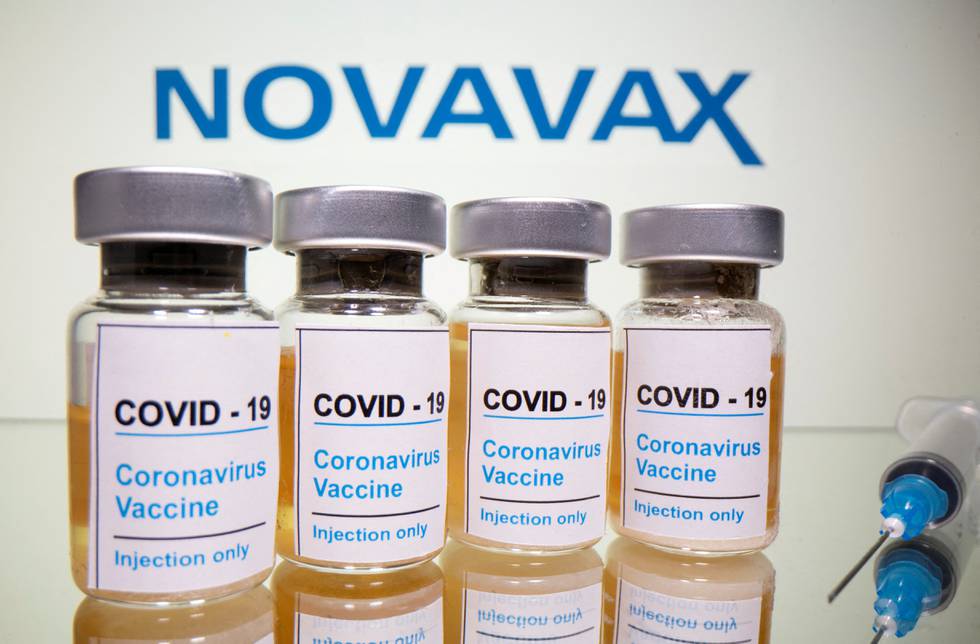 Bildet er av fire små flasker merket med at det er vaksine mot korona. På høyre side ligger det en sprøyte. I bakgrunnen står "Novavax" med blå bokstaver på en lys vegg.