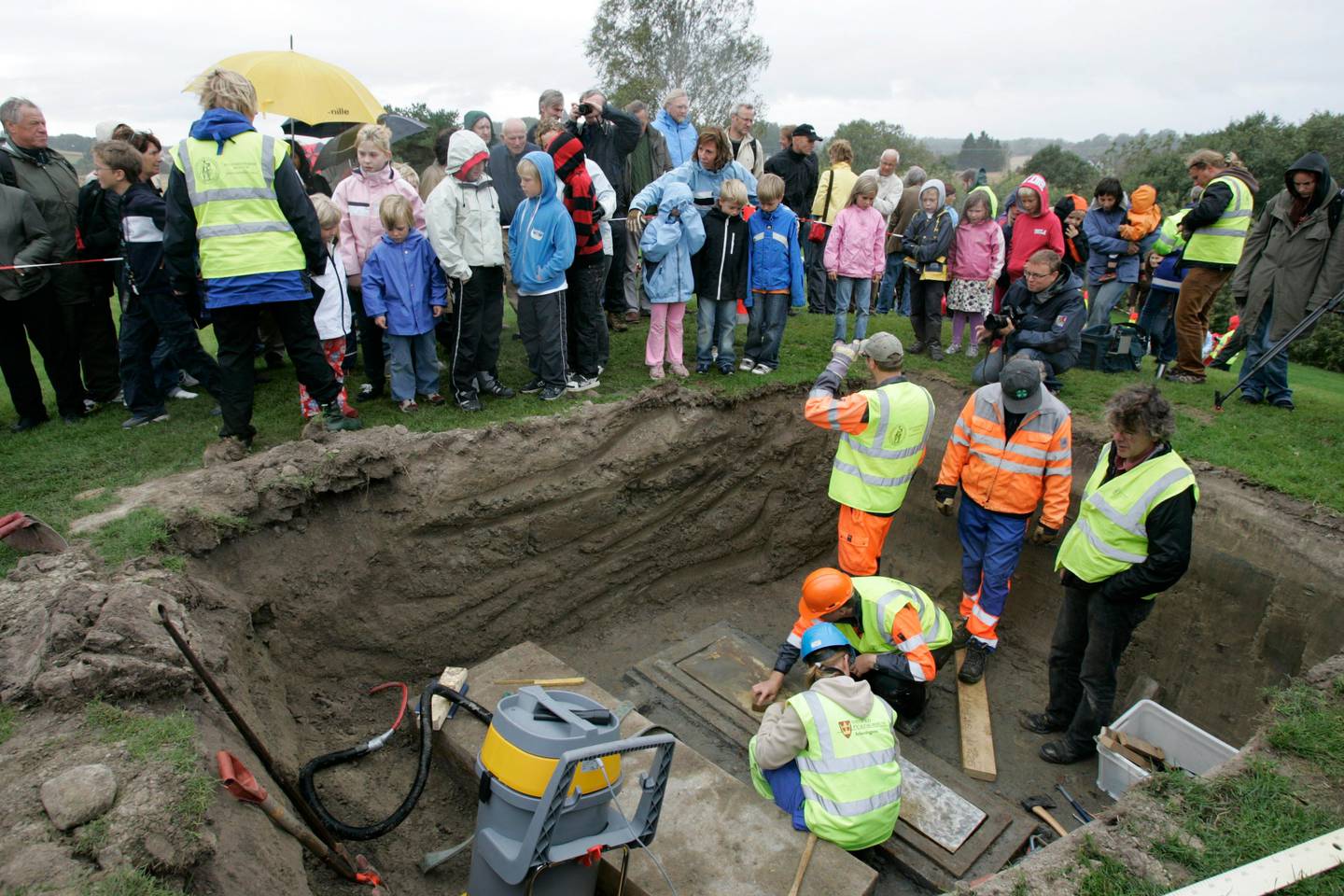 Bildet er av flere personer som står nede i en viking-grav som de har åpnet. Det står barn på kanten av hullet som er gravd ut. De har funnet en kiste.