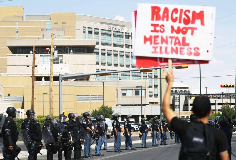 Bildet viser politi med hjelmer og skjold utenfor sykehuset i El Paso. I forgrunnen står en mann med en plakat mot Trump.