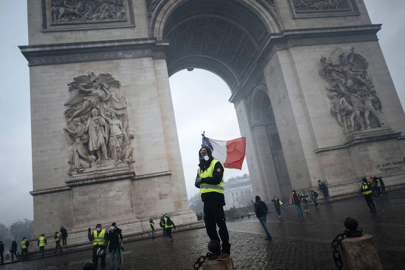 Bildet viser en mann med maske og flagg og gul vest. Han står foran Triumfbuen i Paris i Frankrike.