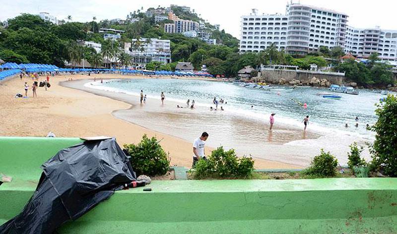 Bildet viser en likpose på en vei ovenfor en strand. Turistene på stranda vet ikke hva som har skjedd på veien over dem. Dette er åstedet for nok et drap i Acapulco i Mexico. 