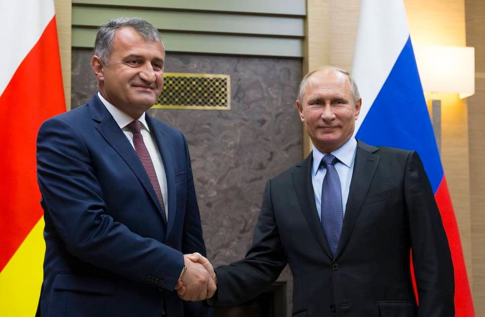 Bildet er av Anatoly Bibilov og Vladimir Putin. De tar hverandre i hendene mens de står foran hvert sitt flagg. Et sør-ossetisk, og et russisk. Bildet er fra 2017. Arkivfoto: van Sekretarev / AP / NTB