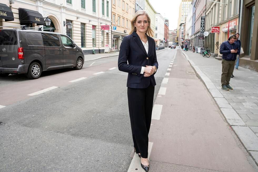Justisminister Emilie Enger Mehl står på gaten med foldede hender. Hun ser mot kamera og har på seg dress. Bak henne står en bil parkert ved fortauet. Det er en svart varebil.