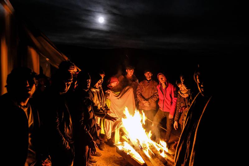 Bildet er av migranter og flyktninger som står rundt et bål. Det eer natta og månen er synlig på himmelen.