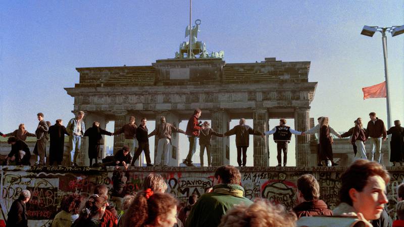 Bildet viser folk som står på muren hånd i hånd. I bakgrunnen er Brandenburger Tor.