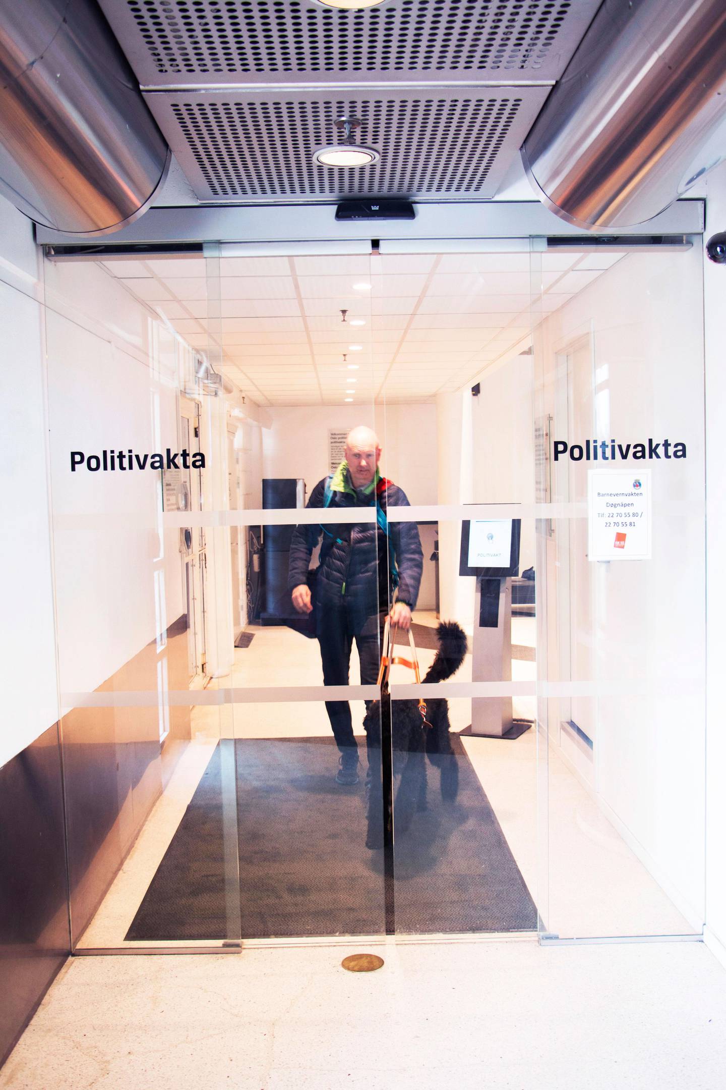 Bildet viser Håkon på vei igjennom en glassdør med skriften «Politivakten». Foran ham går førerhunden Rames. Håkon jobber på Politihuset i Oslo. Der tar han imot tips via telefon og e-post. 