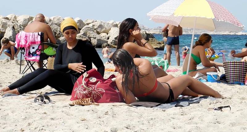 Bildet er av folk på en sandstrand. I fokus på bildet er tre kvinner. To har bikini. Den tredje har burkini. Det er et heldekkende bade-antrekk som kan beskrives som en tynn bukse til anklene, som også sitter sammen med en tynn genser som går til håndleddene. Den dekker også håret på hodet.