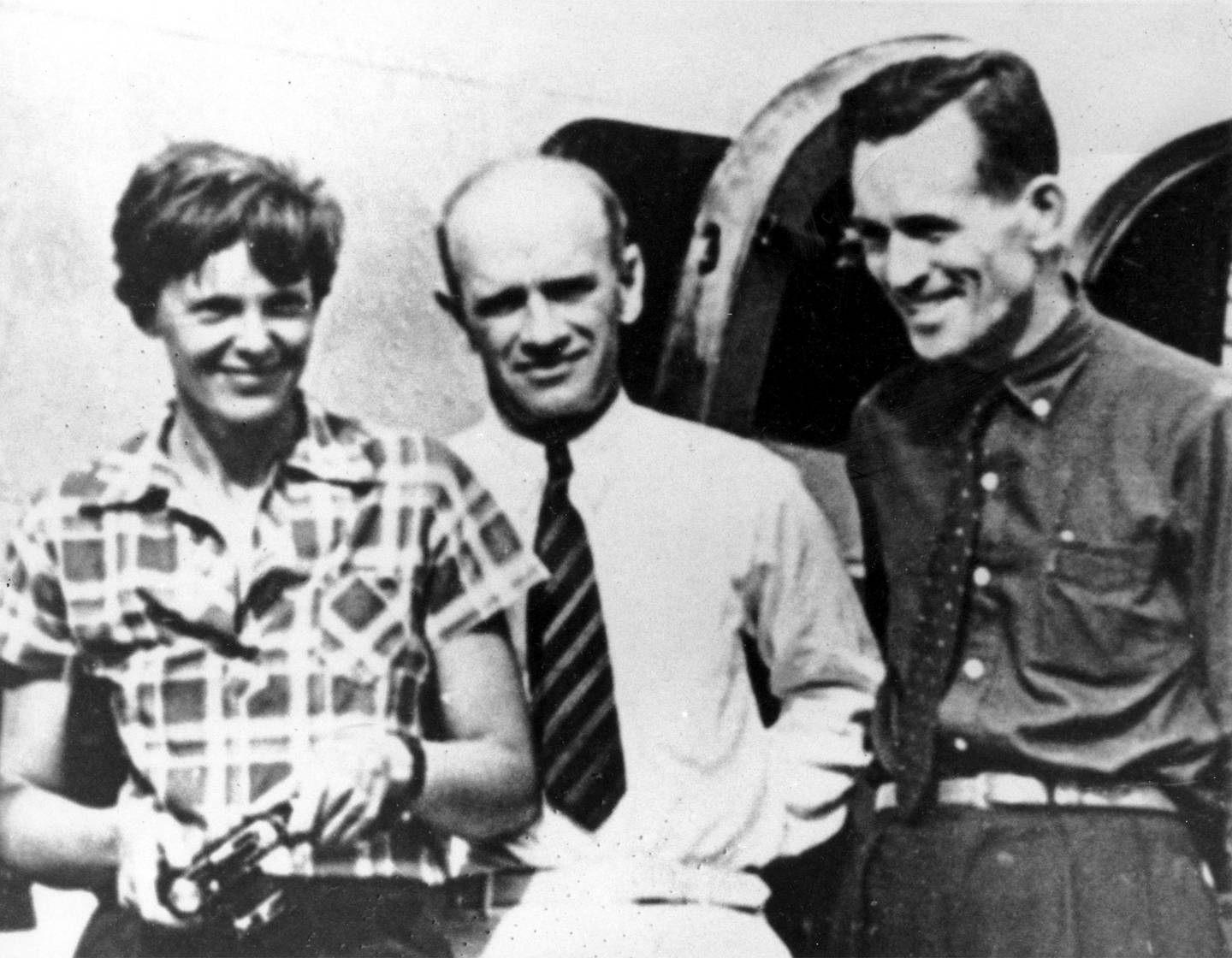Bildet er et svart-hvitt fotografi. Til venstre står Amelia Earhart, og til høyre står Fred Noonan. I midten står en mann med hvit skjorte og mørkt slips. De står foran et fly. Arkivfoto: AP / NTB