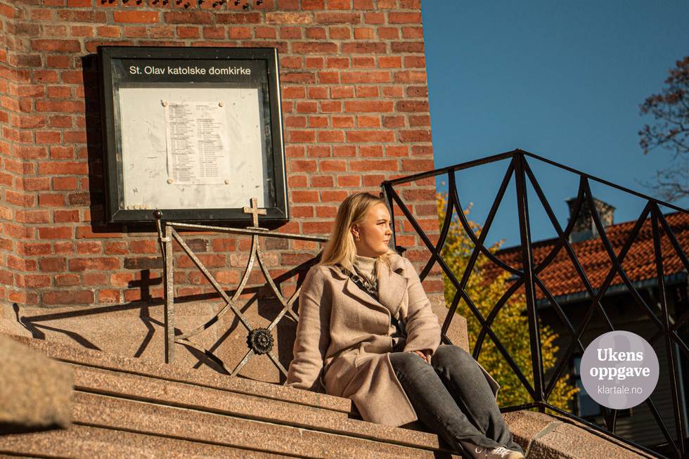Bildet er av en ung dame i en lys frakk. Hun sitter foran en kirke i rød mur.