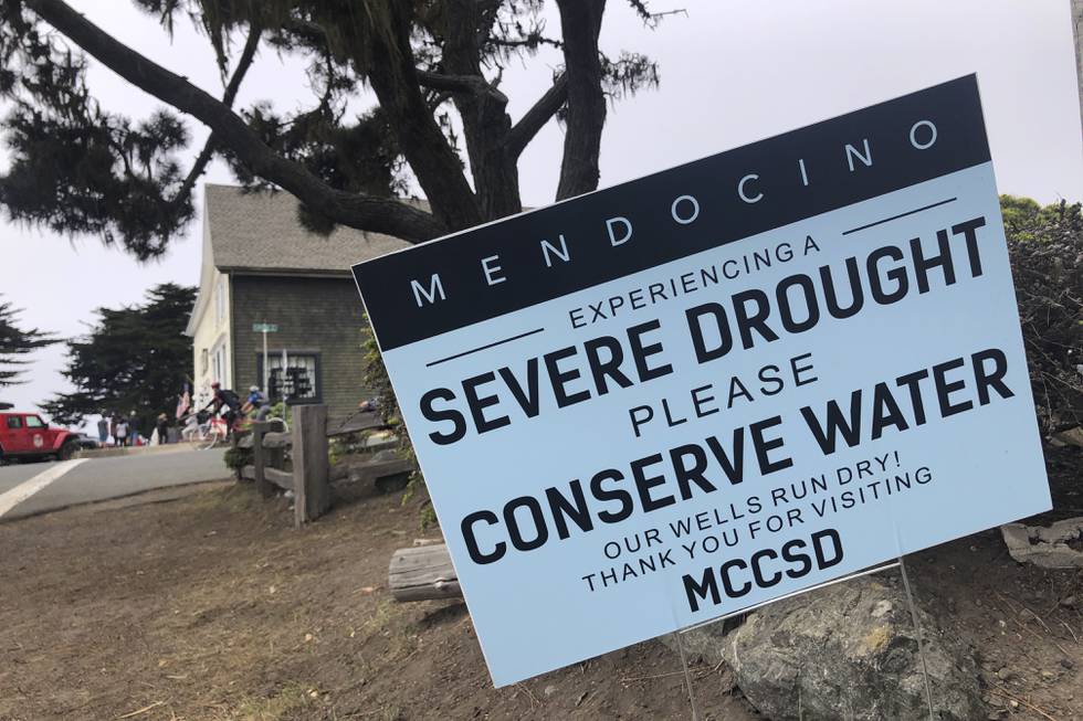 Bildet er av et skilt i byen Mendocino i California.  Det står at folk blir bedt om å spare vann som følge av tørken. Det er helt tørt på bakken. Det er et hus i bakgrunnen. Foto: Haven Daley / AP / NTB