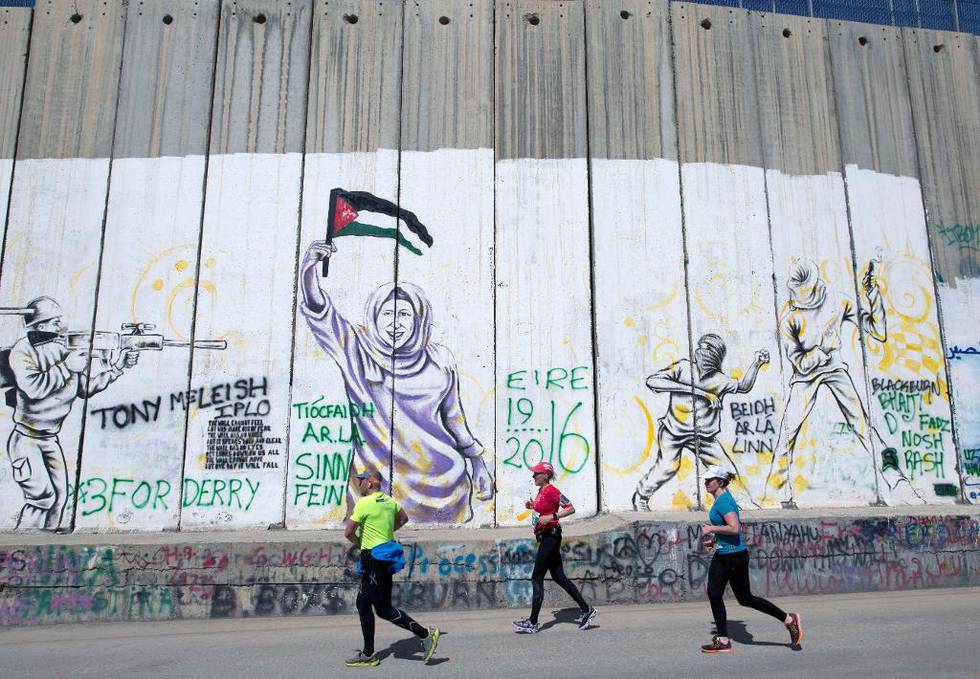 Bildet viser løpere som løper forbi en høy mur. Det er forskjellige tegninger av soldater og kvinner på muren, samt tagging. 