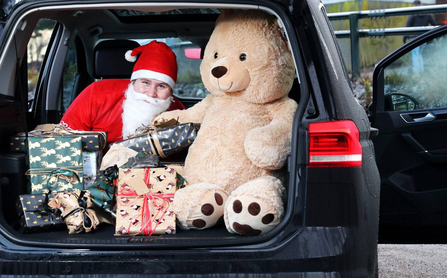 Bildet er av en nisse i bagasjerommet på en bil. Han er gjemt bak gaver og en stor bamse.