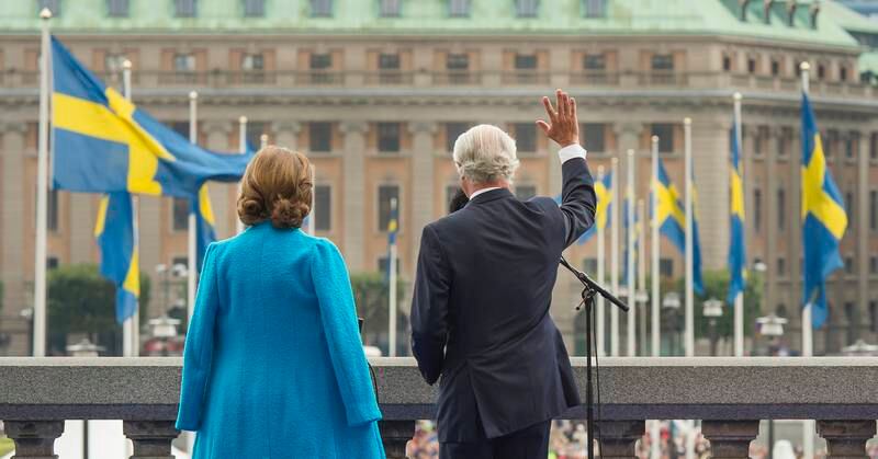 Bildet viser kong Carl Gustaf og dronning Silvia som vinker til folk. Mange flagg vaier foran dem.