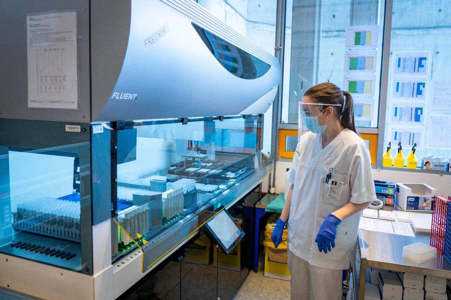 Bildet er av en laboratorie-ansatt som står ved en maskin. Den blir brukt til å sjekke prøver for korona. Det er mange prøver inne i maskinen.
