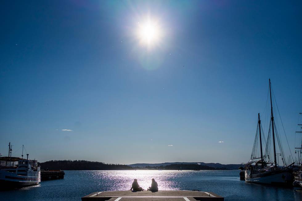 Bildet er av to personer som sitter på ei brygge i sola. Det er seilbåter på sidene.