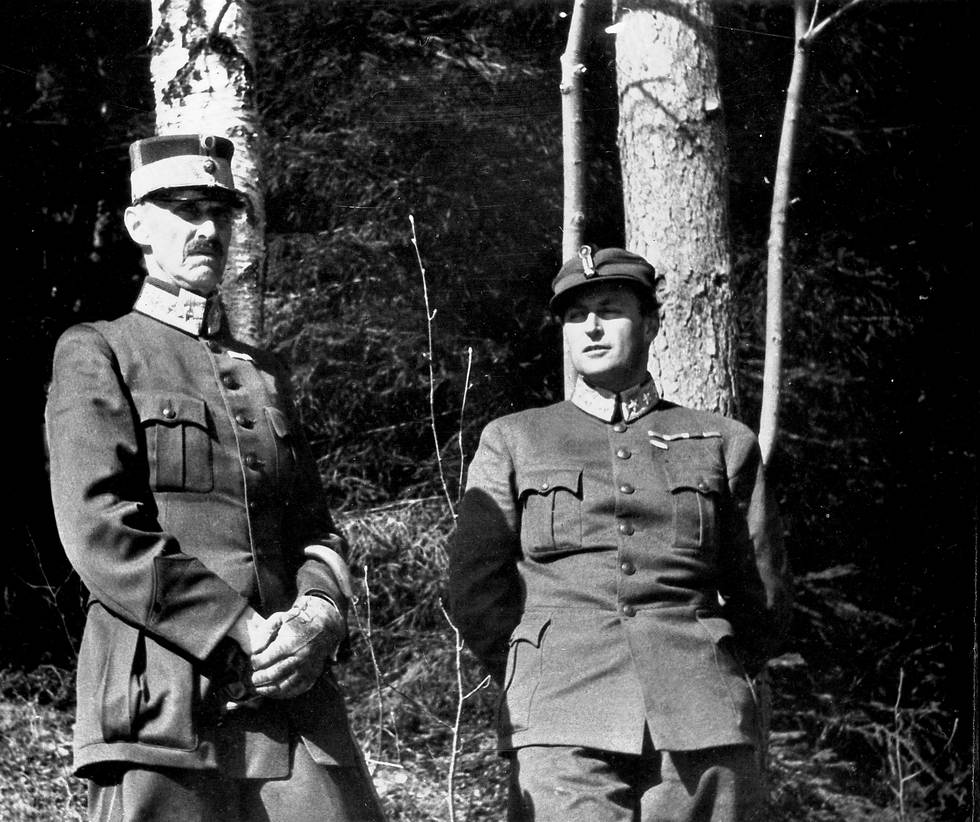 Bildet er av Kong Haakon og kronprins Olav (til høyre). Bildet er tatt foran noen trær. Begge har på militære uniformer. Bildet er i sort/hvitt.