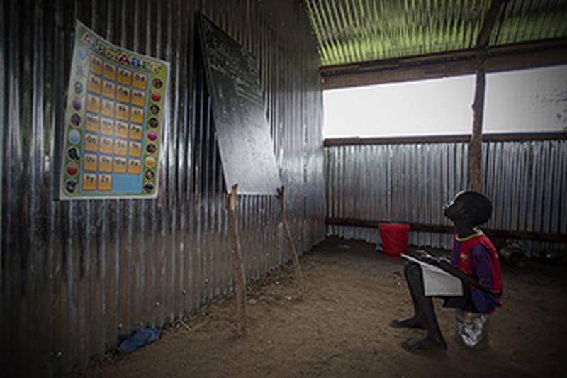 Bildet viser en gutt som sitter på en blikktønne og ser på en tavle. Han går på skole i Sør-Sudan.