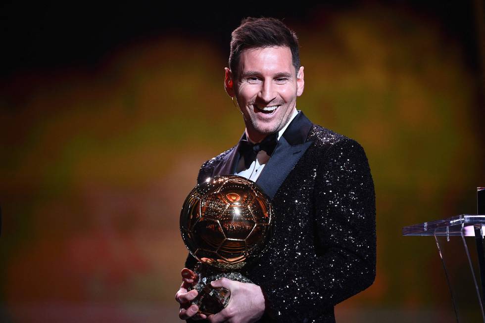 Bildet er av Lionel Messi som holder Gullballen for 2021. Det er et trofé, formet som en ball. Han har på seg svart smoking med paljetter som glitrer i lyset. Han smiler.