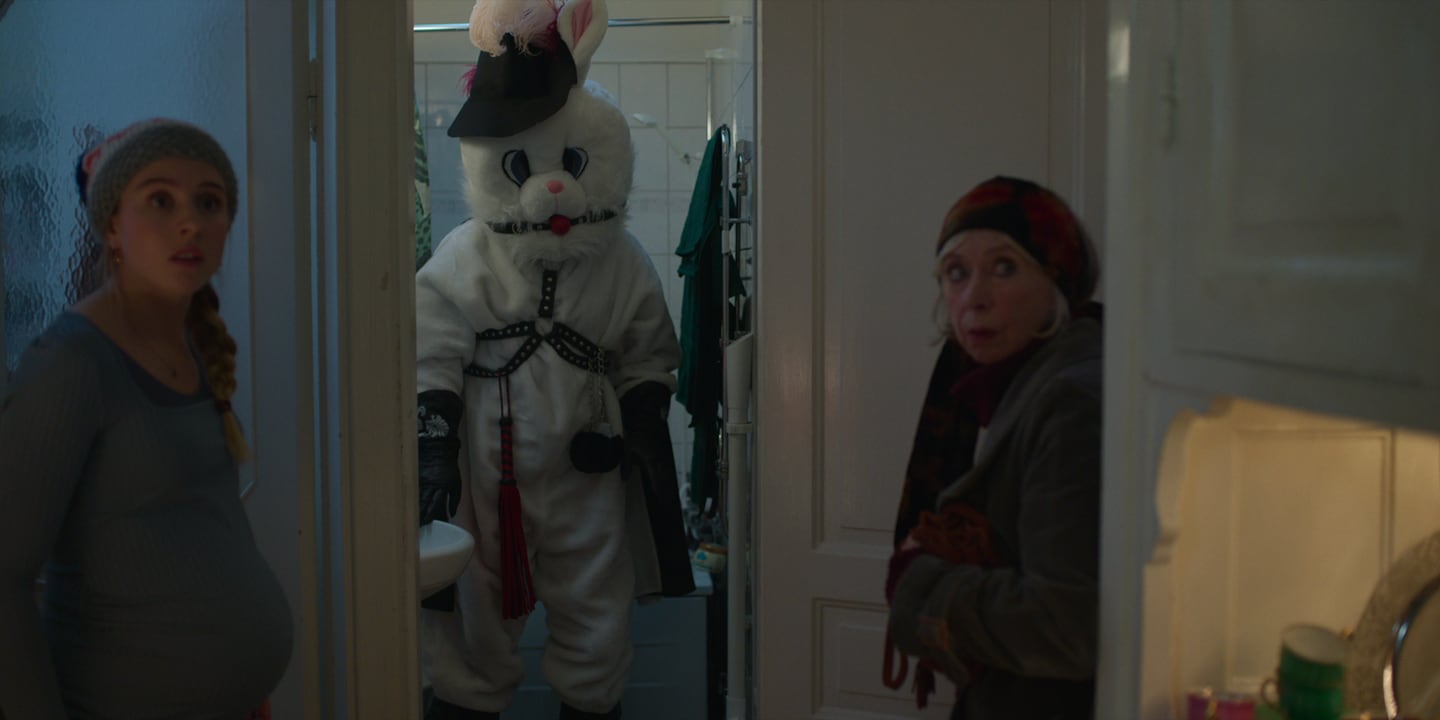 Bildet er av to damer som står på hver sin side av en døråpning. I åpningen står en person i kaninkostyme, med BDSM-detaljer.