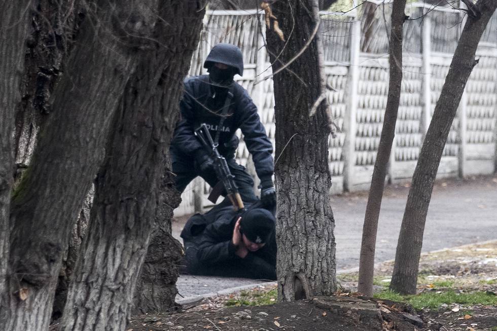 Bildet er av en væpnet politimann med maske og hjelm som sikter et automatvåpen i nakken på en demonstrant som ligger på bakken med ansiktet ned. Det er mulig å se dem mellom noen trær. Foto: Vasily Krestyaninov / AFP / NTB