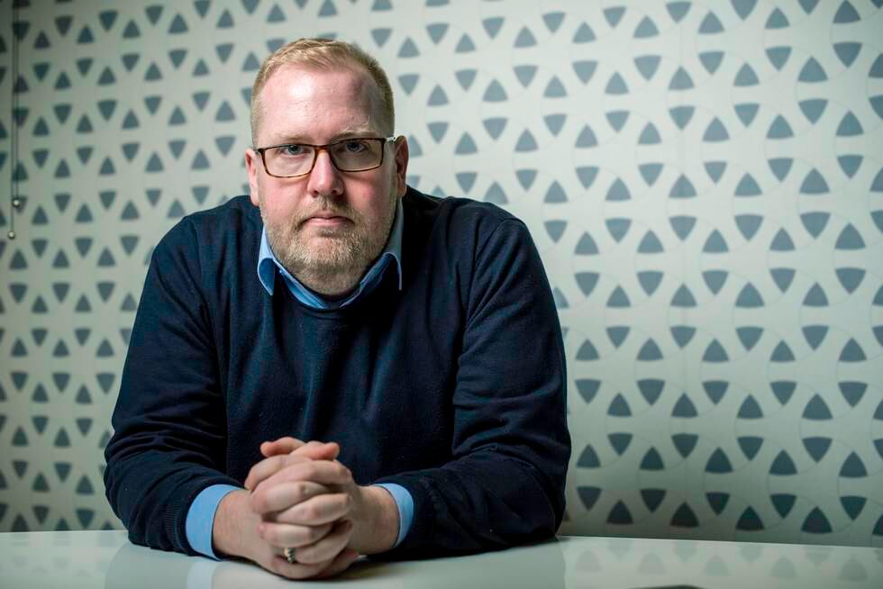 Bildet er av Tor Bernhard Slaathaug. Han sitter med foldede hender. Han har på en mørkeblå genser over en lyseblå skjorte. Han sitter ved et bord, foran en hvit vegg med gråblått mønster av trekanter. Han har helskjegg og briller med mørk innfatning.