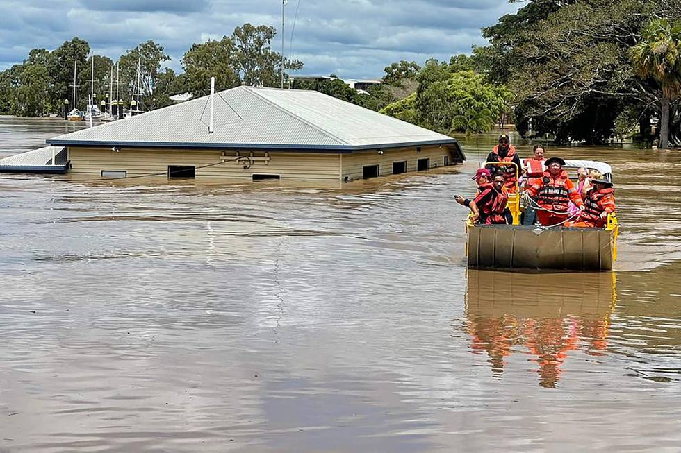 Bildet er folk ombord i en båt. Den er ute på vannet under en flom. De passerer et hus som har vann nesten opp til taket. Bildet er fra flommen i Maryborough i Queensland i Australia.