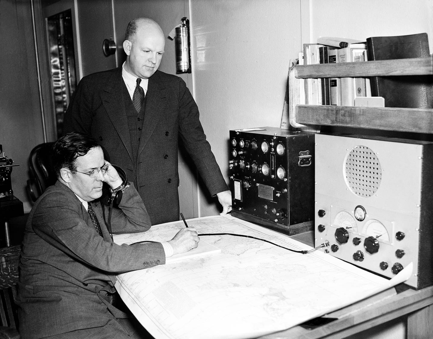 Bildet er av to menn. En av dem står, og den andre sitter. George Palmer Putnam er ektemannen til Earhart, og står mens han ser på et stort kart. Det ligger på et bord hos kystvakten. Arkivfoto: NTB