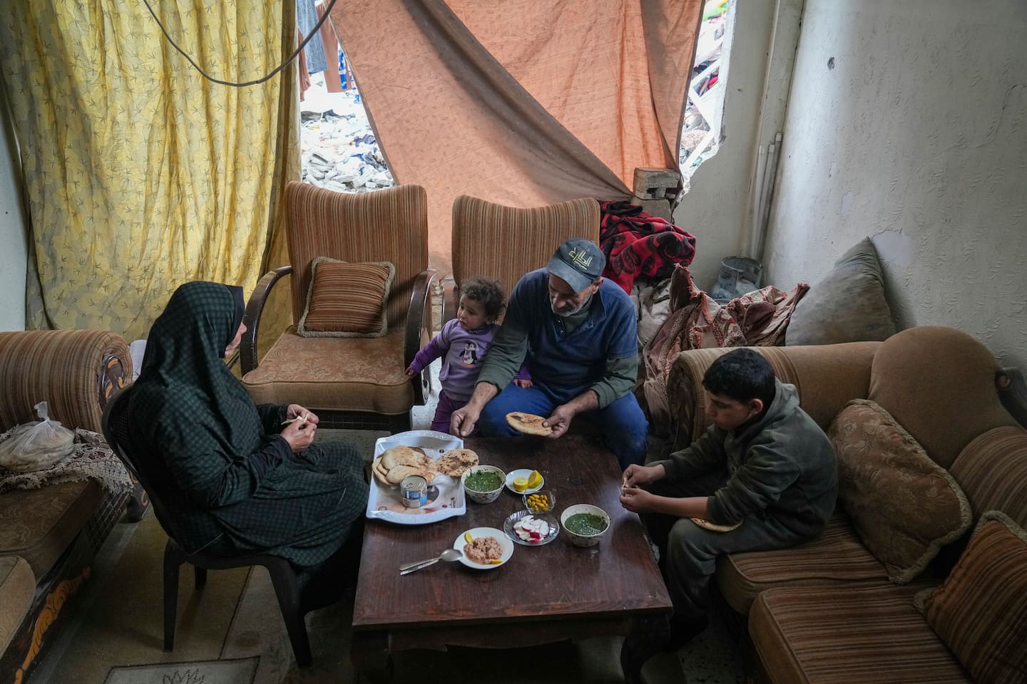 Khadra Abu Libda og mannen Khalid Nassar spiser lunsj med barnebarna sine. Et laken skiller stuen deres fra ruinene utenfor. Foto: Abdel Kareem Hana / AP / NTB