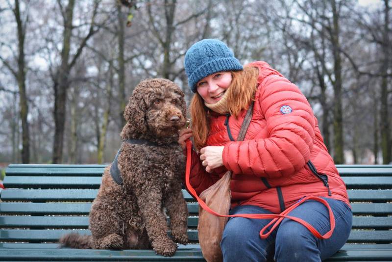 Bildet er av Grete Moene og hunden Ella. Det er en hund med brun og krøllete pels. De sitter på en benk i parken.