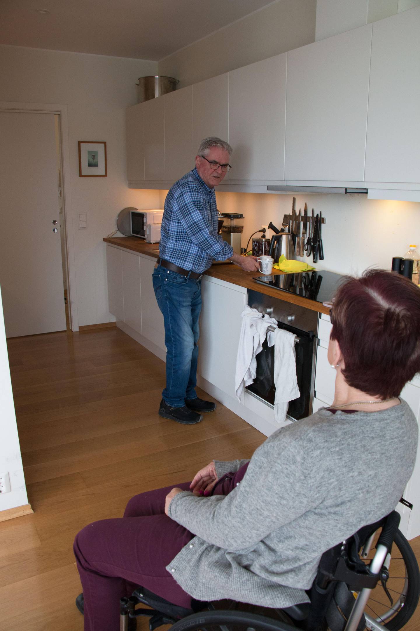Bildet viser Ågot som sitter i rullestol og ser på at Yngve lager kaffe på kjøkkenet.