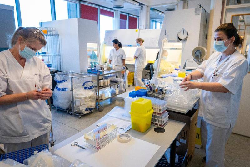 Bildet er av laboratorie-ansatte på Ullevål sykehus. I forgrunnen står to stykker langs en benk med mange prøver. I bakgrunnen står to andre ved en tralle med prøver. En femte sitter ved en maskin og sjekker prøvene.